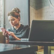 Young businesswoman sitting in cafe at table near window and using smartphone. On desk is laptop and cup of coffee. Girl working, learning online. Social media, network. Online marketing, education.