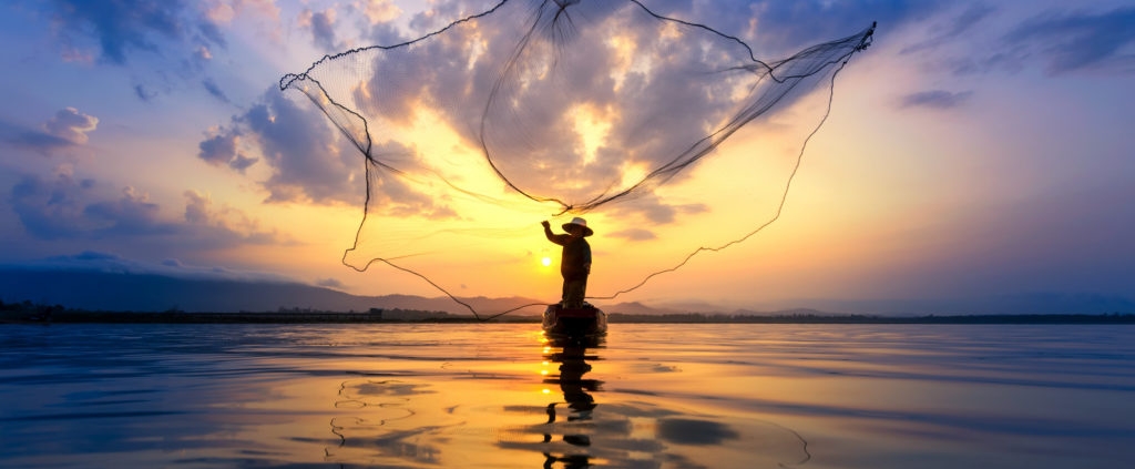 fisherman on wooden boat casting a net for catching freshwater fish in nature river in the early morning before sunrise