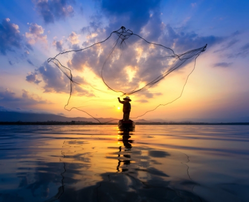 fisherman on wooden boat casting a net for catching freshwater fish in nature river in the early morning before sunrise